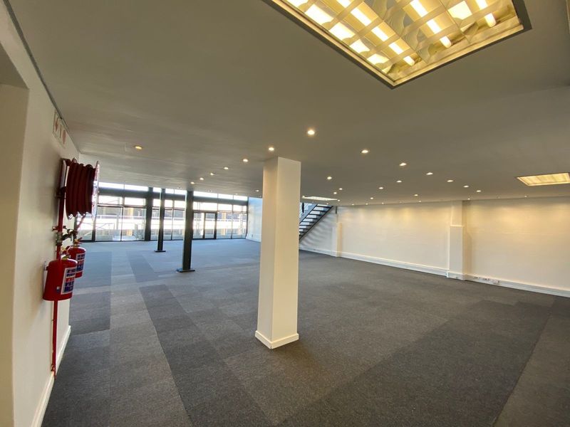 A spacious, open-plan office area featuring neutral carpeting, recessed lighting, structural columns, and large windows providing ample natural light.
