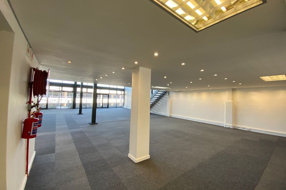 A spacious, open-plan office area featuring neutral carpeting, recessed lighting, structural columns, and large windows providing ample natural light.