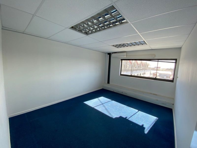 An empty office space featuring a suspended ceiling with recessed lighting, blue carpeting, and a wall-mounted air conditioning unit.