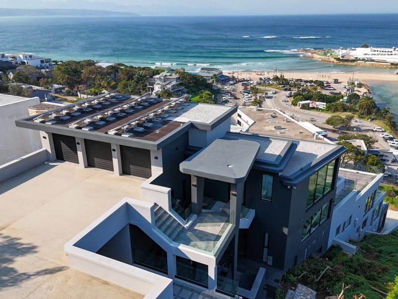 Aerial view of a modern multi-level coastal residence featuring a triple garage, solar panel array, and expansive views of the ocean and beach.
