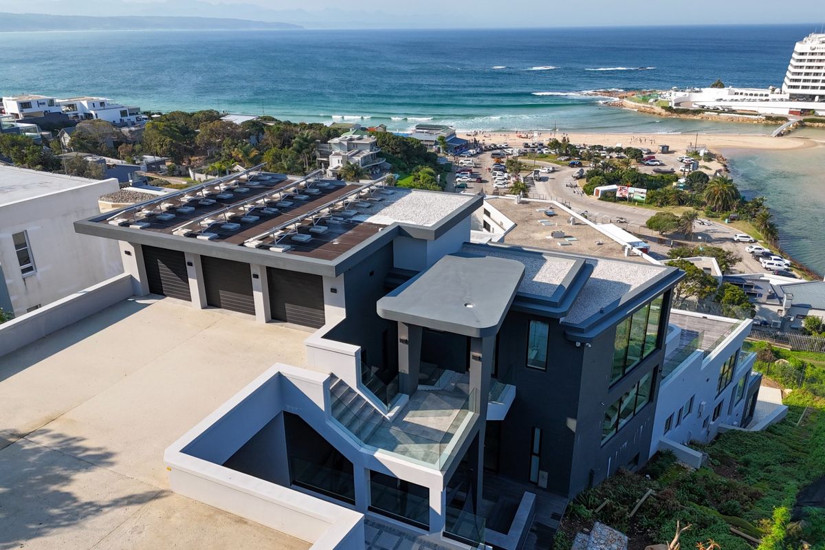 Aerial view of a modern multi-level coastal residence featuring a triple garage, solar panel array, and expansive views of the ocean and beach.