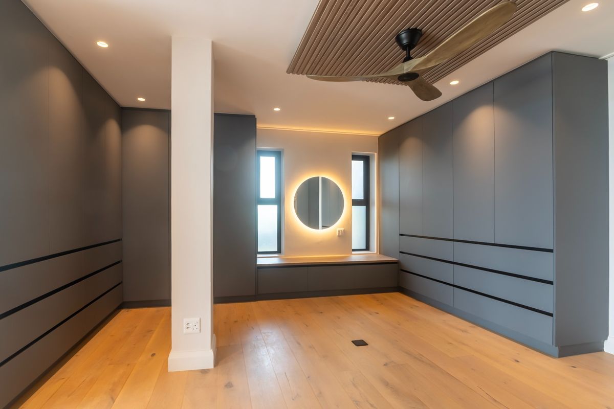 Modern bedroom featuring extensive built-in cabinetry, timber flooring, a decorative timber ceiling panel with a fan, and a backlit vanity mirror.