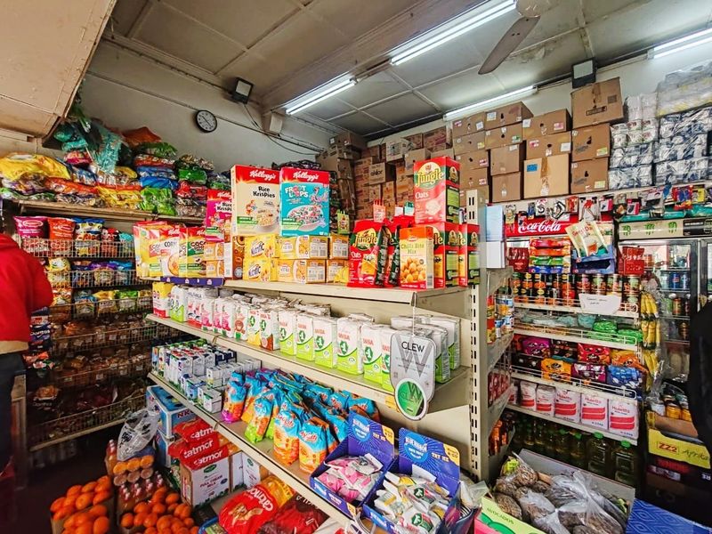 Interior view of a retail store featuring metal shelving units stocked with various grocery products, overhead fluorescent lighting, and a ceiling fan.