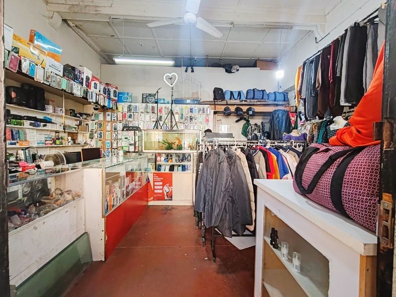 A retail shop interior featuring built-in shelving units, fluorescent lighting, and a ceiling fan, currently utilized for clothing and electronics display.