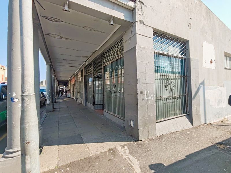 Street-level view of a retail storefront featuring a covered walkway, security grilles, and large display windows in need of exterior maintenance.