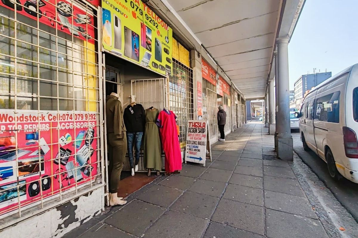 Street-level view of a retail storefront featuring a covered walkway, security gates, and signage along a paved sidewalk.