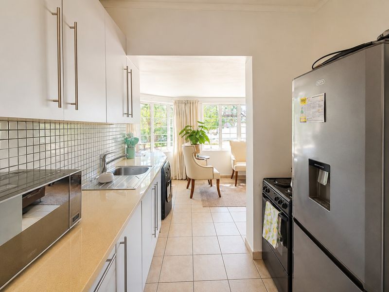 A functional kitchen featuring white cabinetry, tiled backsplash, laminate countertops, and a stainless steel sink, leading into a bright adjacent living area.