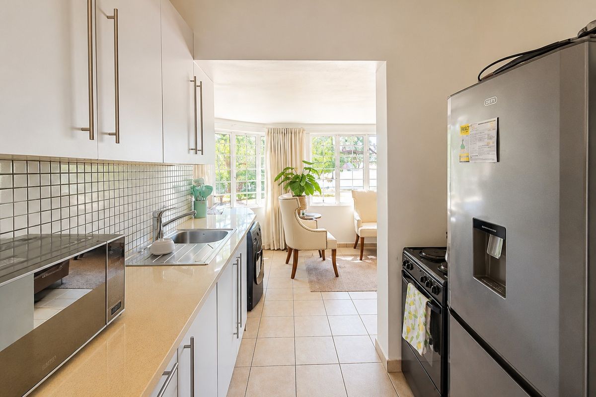 A functional kitchen featuring white cabinetry, tiled backsplash, laminate countertops, and a stainless steel sink, leading into a bright adjacent living area.