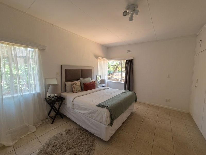 A well-lit bedroom featuring neutral tiled flooring, built-in cupboards, and dual aspect windows providing natural light.