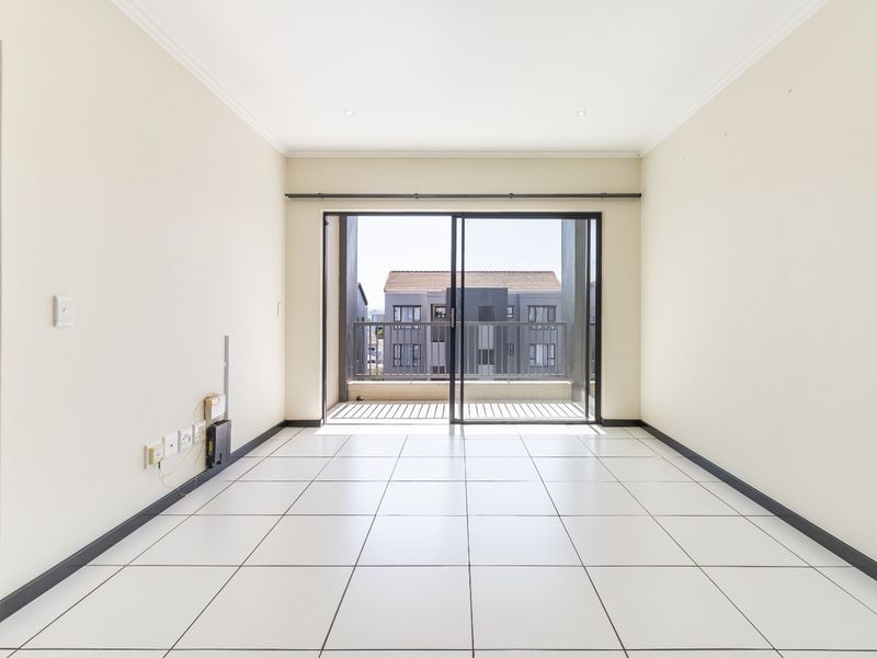 A bright, empty living room featuring white tiled flooring, recessed lighting, and a sliding glass door leading to a private balcony.