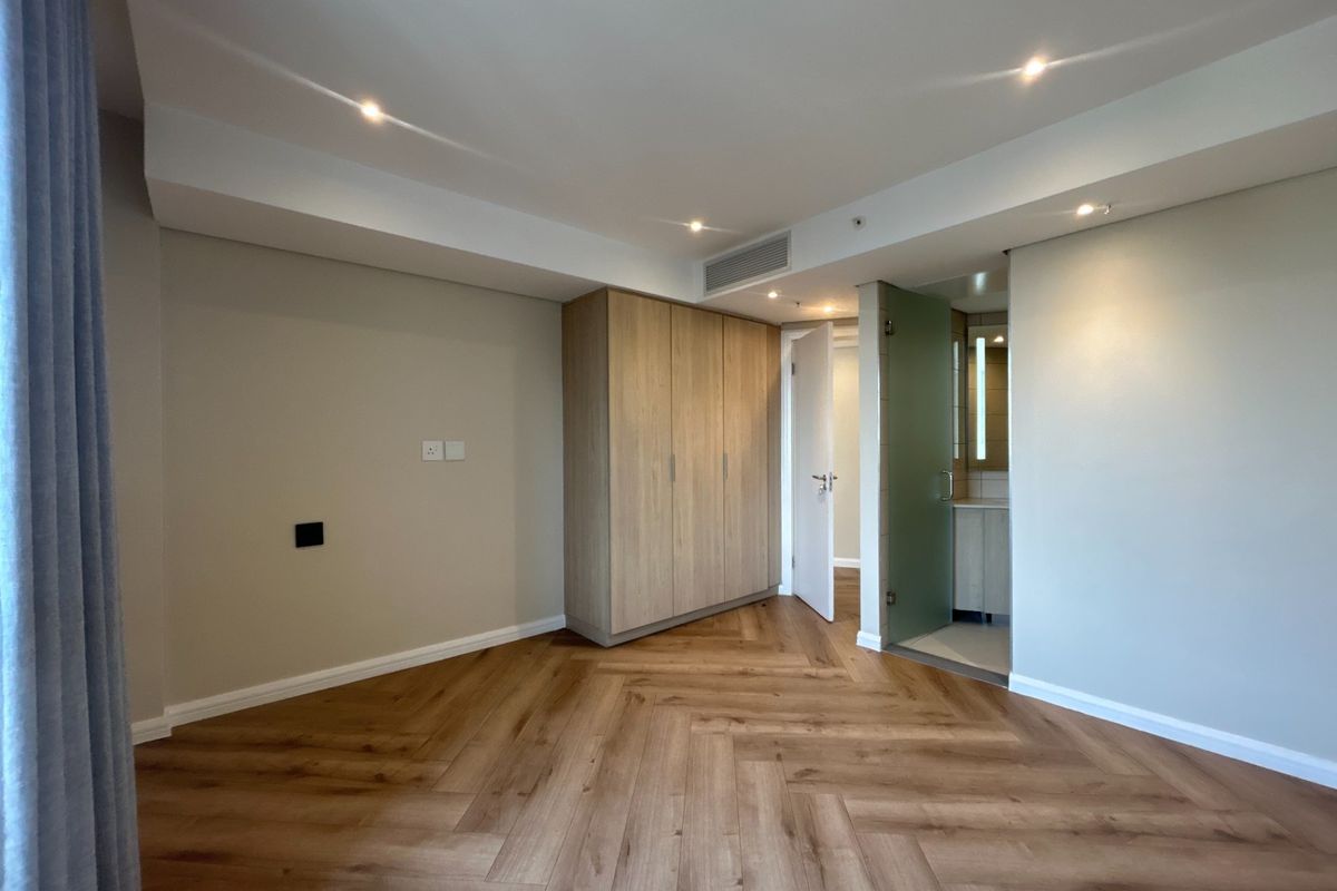 A modern, vacant bedroom featuring high-quality herringbone timber flooring, built-in cabinetry, recessed lighting, and direct access to an en-suite bathroom.