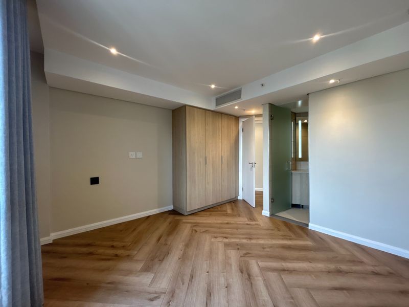 A modern, vacant bedroom featuring high-quality herringbone timber flooring, built-in cabinetry, recessed lighting, and direct access to an en-suite bathroom.