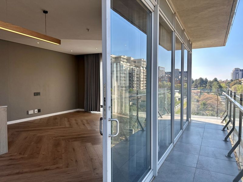 Modern open-plan living area featuring herringbone timber flooring, floor-to-ceiling glass doors, and a spacious balcony with glass balustrades offering urban views.