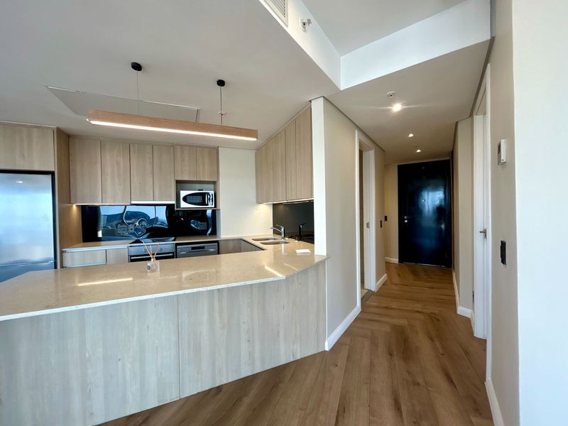 Modern open-plan kitchen featuring light timber cabinetry, stone benchtops, integrated appliances, and elegant herringbone timber flooring in a well-maintained apartment setting.