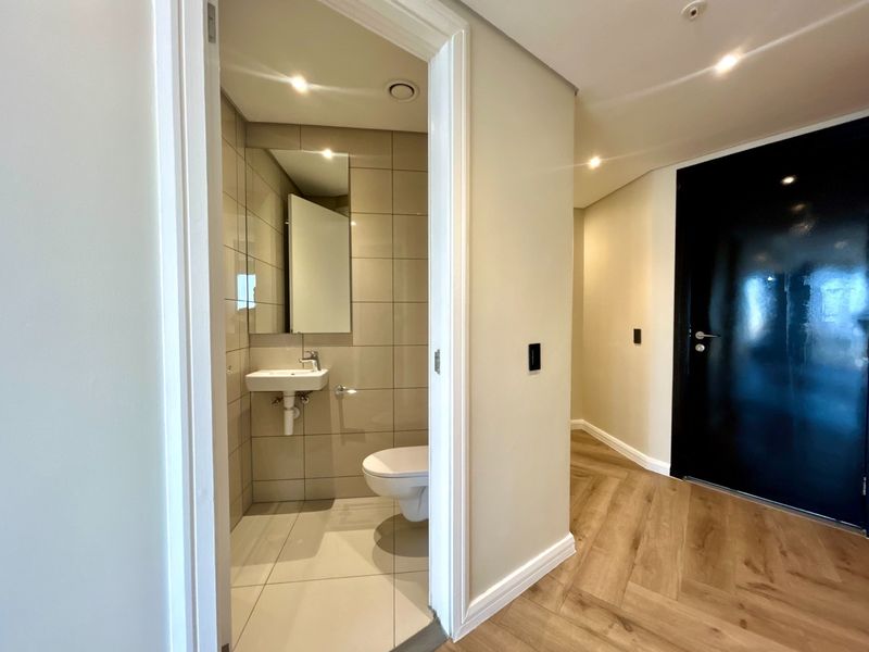 A modern guest bathroom featuring a wall-mounted toilet and basin, complemented by neutral wall tiles and adjacent wood-look laminate flooring in the hallway.