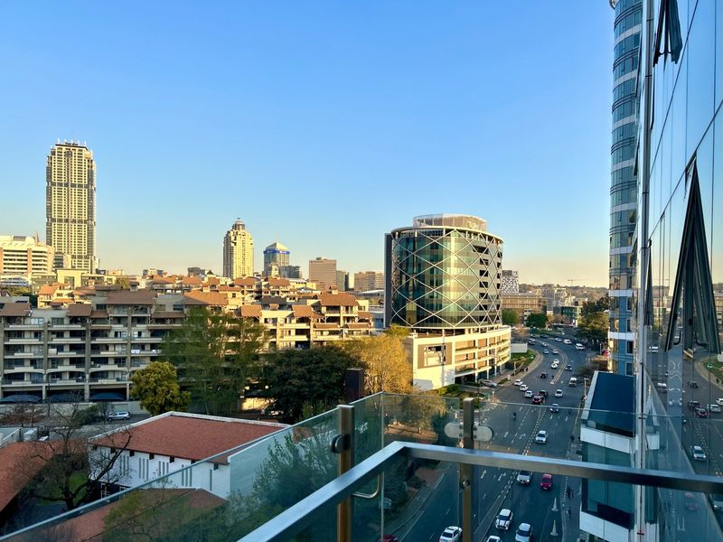Elevated urban view overlooking the city skyline and surrounding streets, captured from a modern apartment balcony with a glass balustrade.