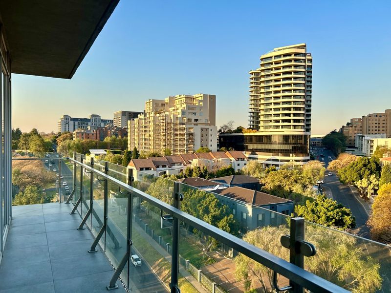 A modern balcony featuring a sleek glass balustrade and tiled flooring, offering an expansive elevated view of the surrounding urban landscape.