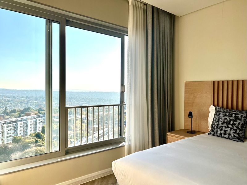 Modern bedroom featuring a large sliding window with expansive city views, integrated wooden headboard, and neutral finishes.