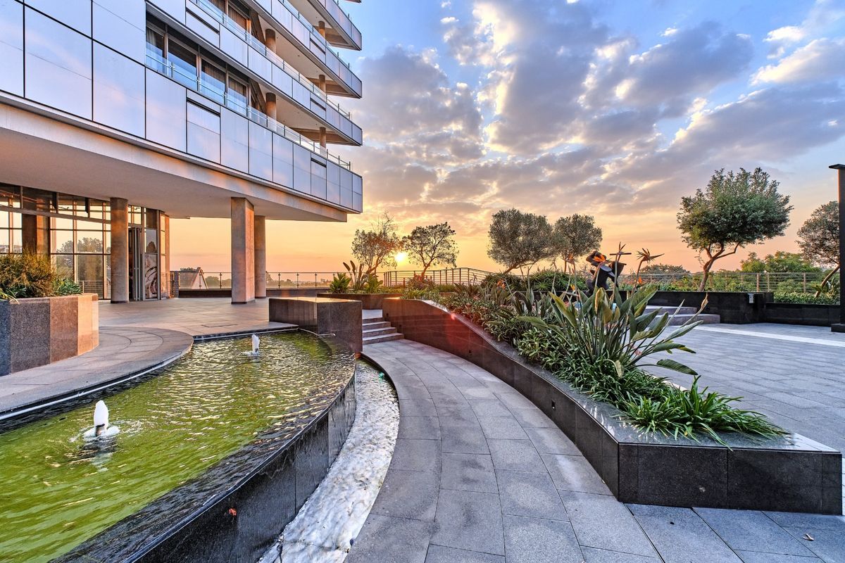 Modern apartment building exterior featuring a landscaped terrace with a stone-paved walkway, water feature, and sunset views.