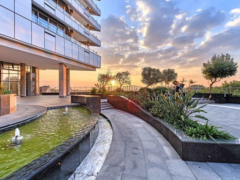 Modern apartment building exterior featuring a landscaped terrace with a stone-paved walkway, water feature, and sunset views.
