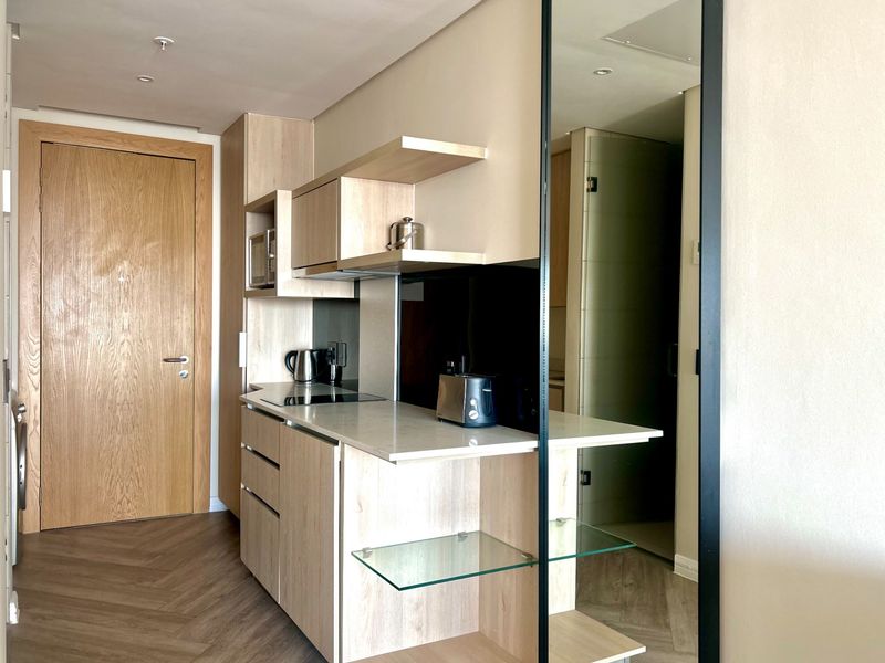 Modern compact kitchen featuring light wood cabinetry, stone countertops, integrated appliances, and stylish herringbone flooring in a clean, contemporary apartment setting.