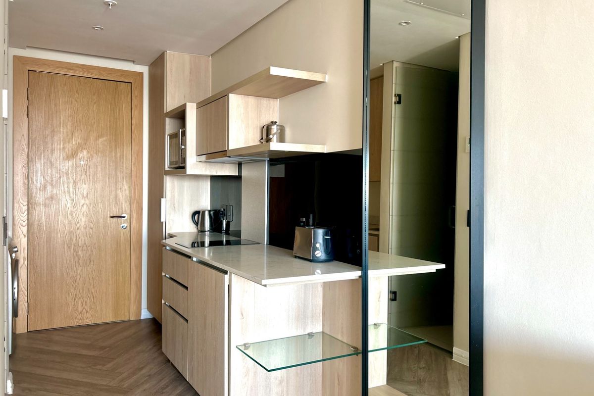 Modern compact kitchen featuring light wood cabinetry, stone countertops, integrated appliances, and stylish herringbone flooring in a clean, contemporary apartment setting.