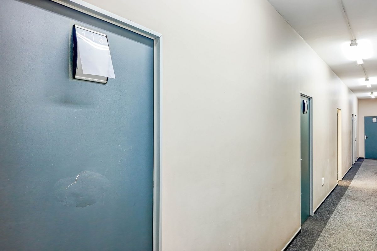 A commercial hallway featuring multiple doors, carpeted flooring, and overhead fluorescent lighting, showing signs of wear and tear.