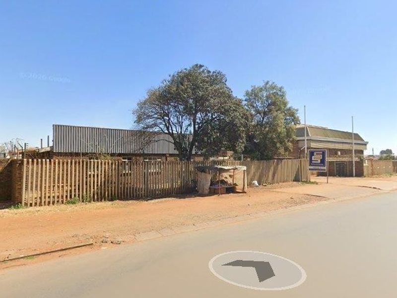 Street view of a commercial property featuring a brick boundary wall, metal fencing, and an exposed steel structure on a dusty roadside.