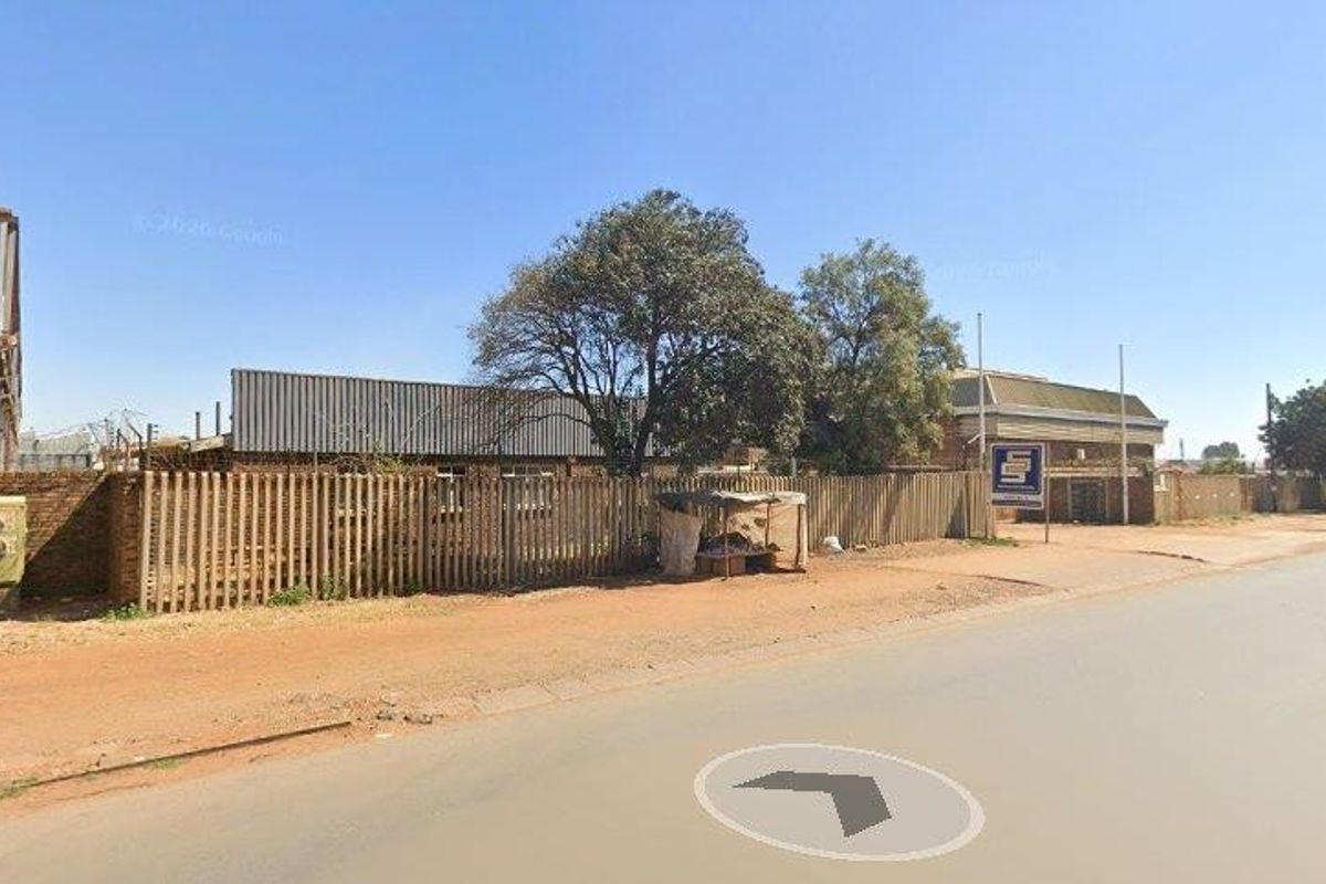 Street view of a commercial property featuring a brick boundary wall, metal fencing, and an exposed steel structure on a dusty roadside.