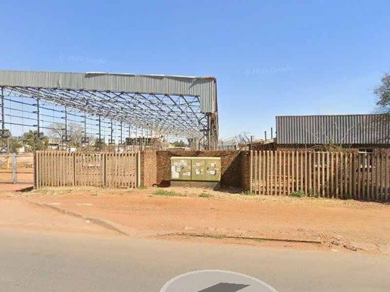Street view of a commercial property featuring a large open steel frame structure, brick perimeter wall, and metal security fencing.