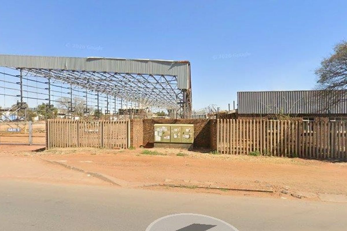Street view of a commercial property featuring a large open steel frame structure, brick perimeter wall, and metal security fencing.