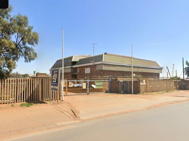Street view of a commercial property featuring a gated entrance, security fencing, and a brick building with a metal roof.