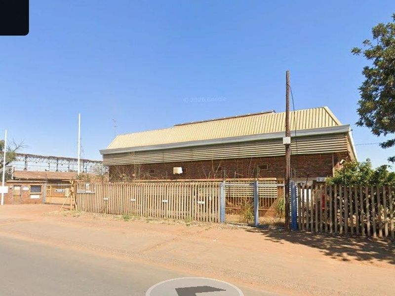 Street view of a commercial property featuring a brick building with a corrugated metal roof, enclosed by metal palisade fencing.