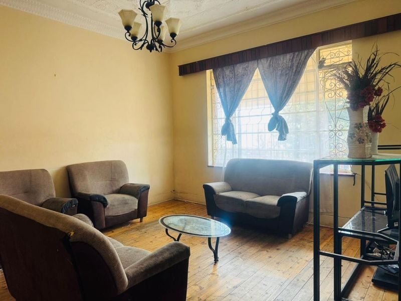 A living room featuring original hardwood flooring, ornate ceiling cornicing, and large windows fitted with security burglar bars.