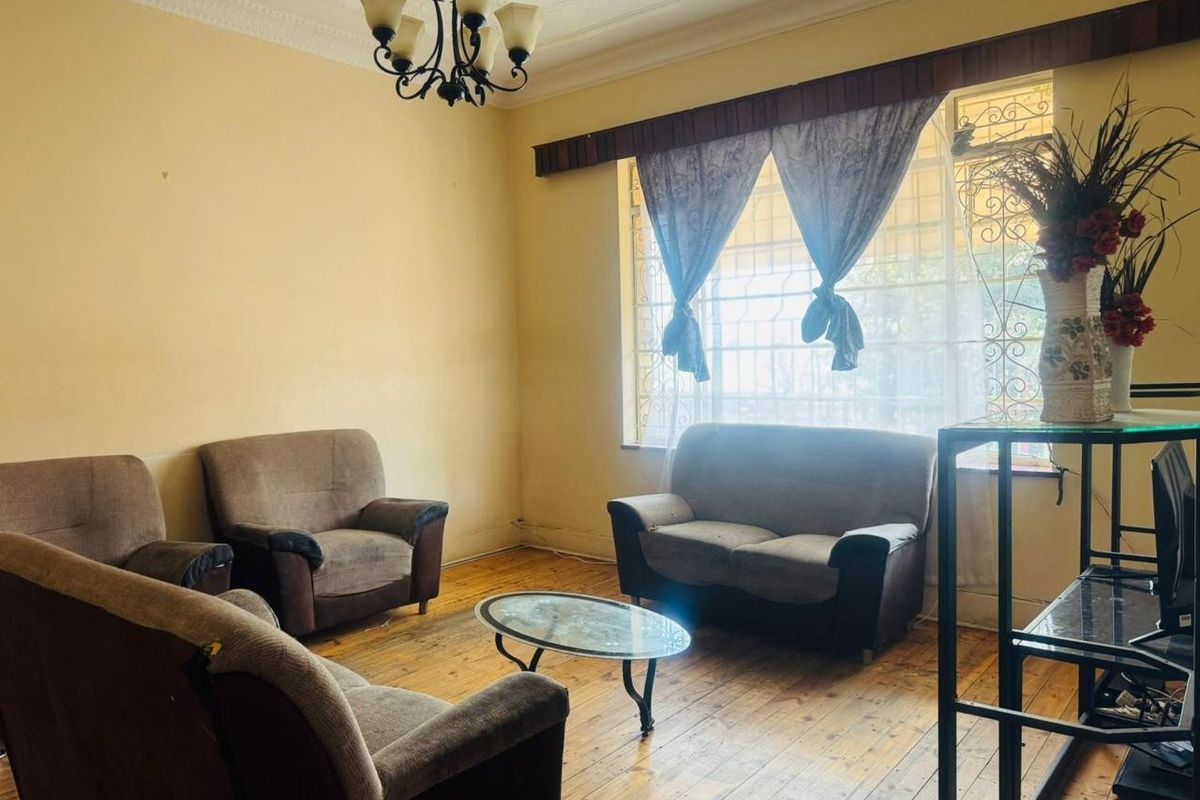 A living room featuring original hardwood flooring, ornate ceiling cornicing, and large windows fitted with security burglar bars.