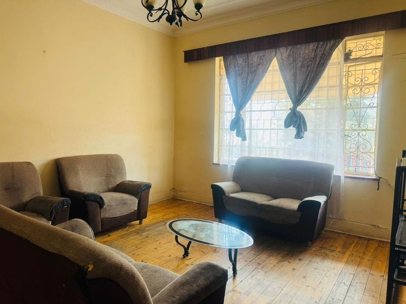A living room featuring original hardwood flooring, decorative ceiling cornicing, and a large window fitted with a wrought iron security gate.