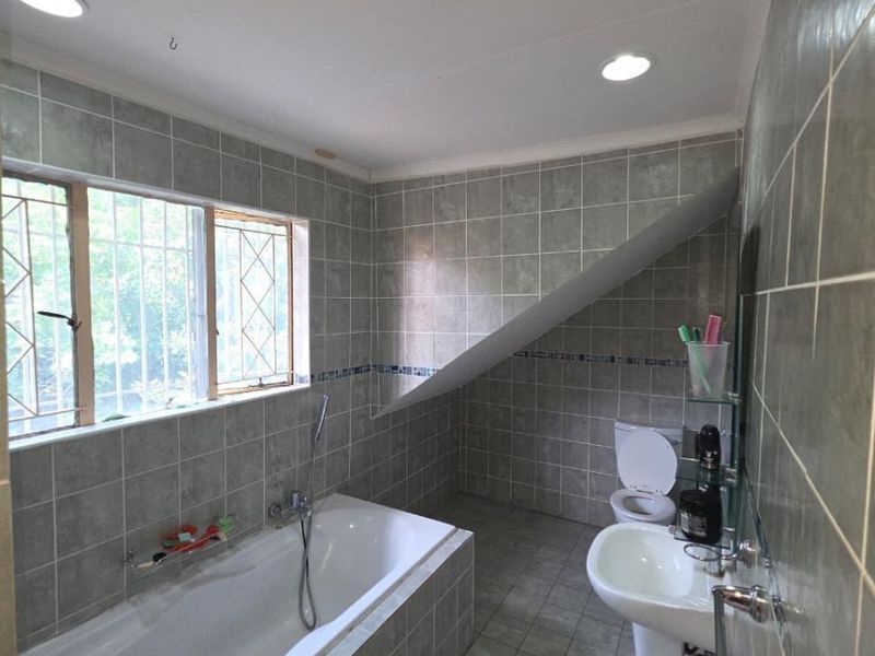 A functional bathroom featuring a built-in bathtub, pedestal basin, toilet, and full-height wall tiling with recessed lighting.
