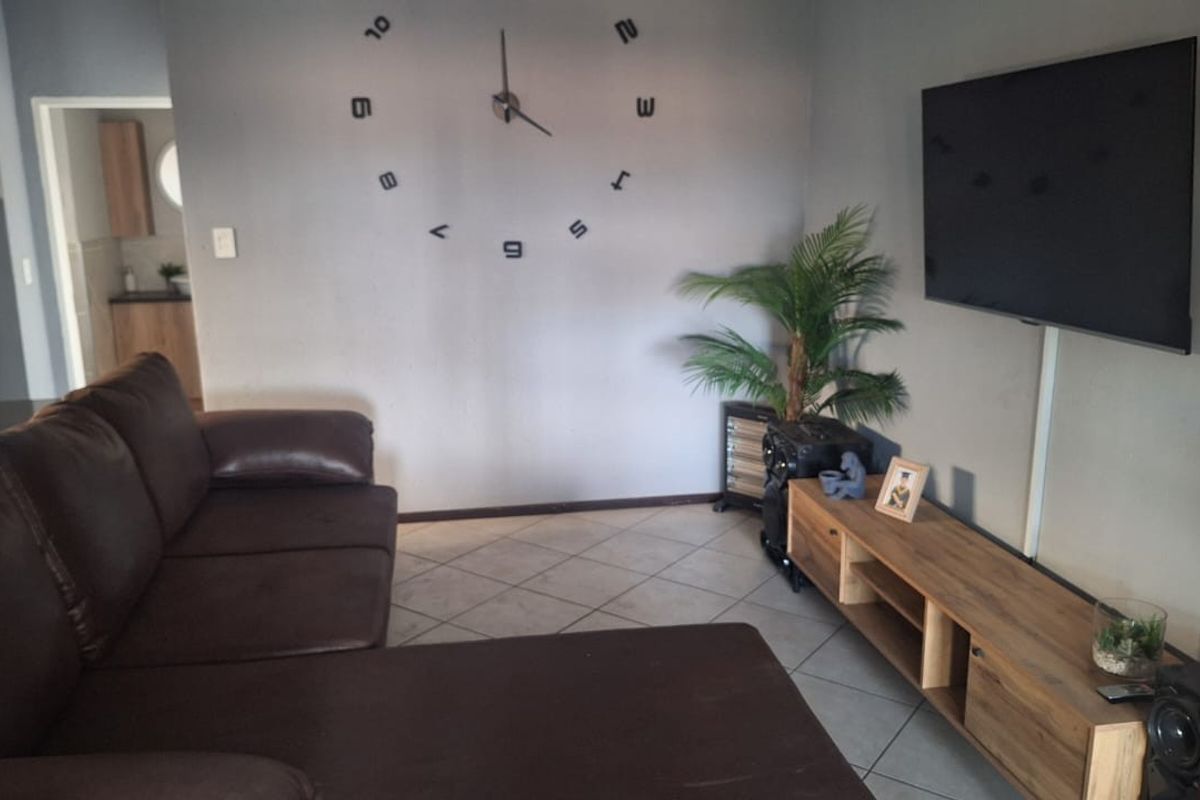 A living room featuring tiled flooring and a wall-mounted television, with a view into an adjacent kitchen area.