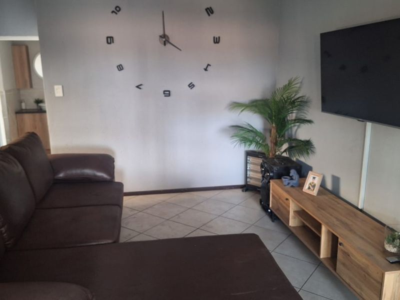 A living room featuring tiled flooring and a wall-mounted television, with a view into an adjacent kitchen area.