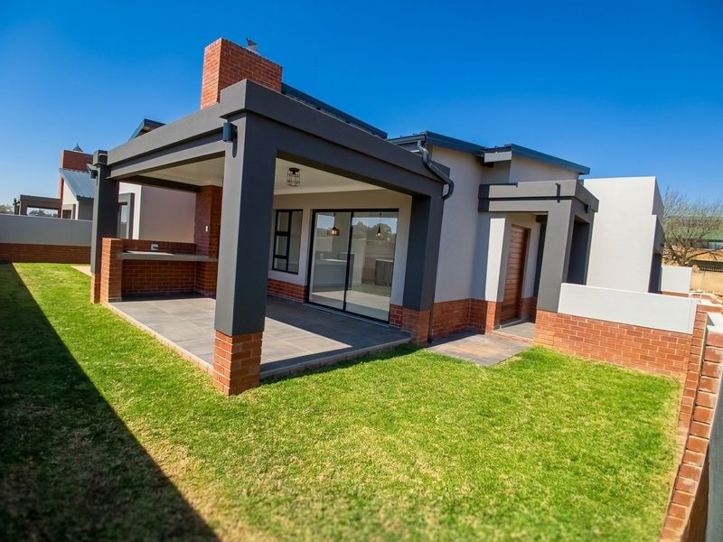 A modern rear exterior view featuring a covered patio with a built-in braai, brick chimney, and sliding glass doors leading to a well-maintained lawn.
