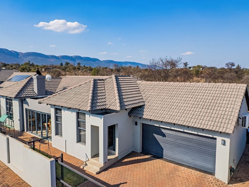 Aerial view of a modern residential property featuring a tiled roof, double garage, paved driveway, and solar panels set against a scenic landscape.