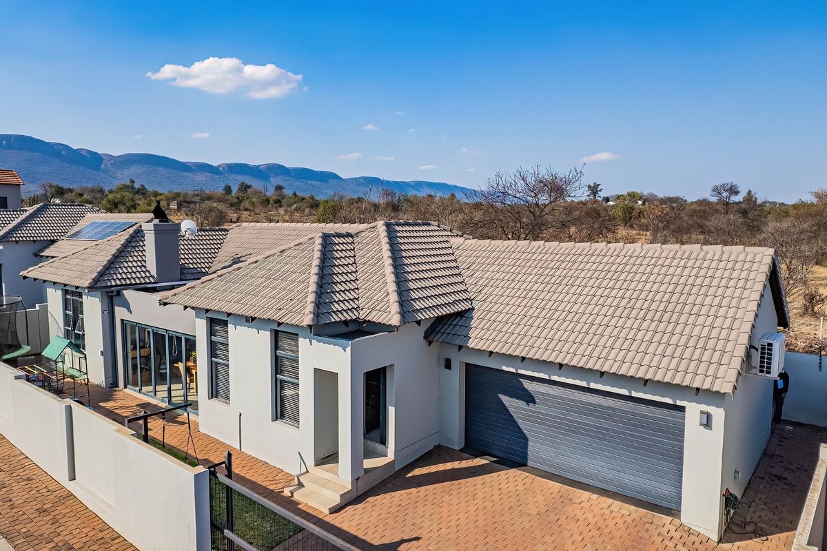 Aerial view of a modern residential property featuring a tiled roof, double garage, paved driveway, and solar panels set against a scenic landscape.