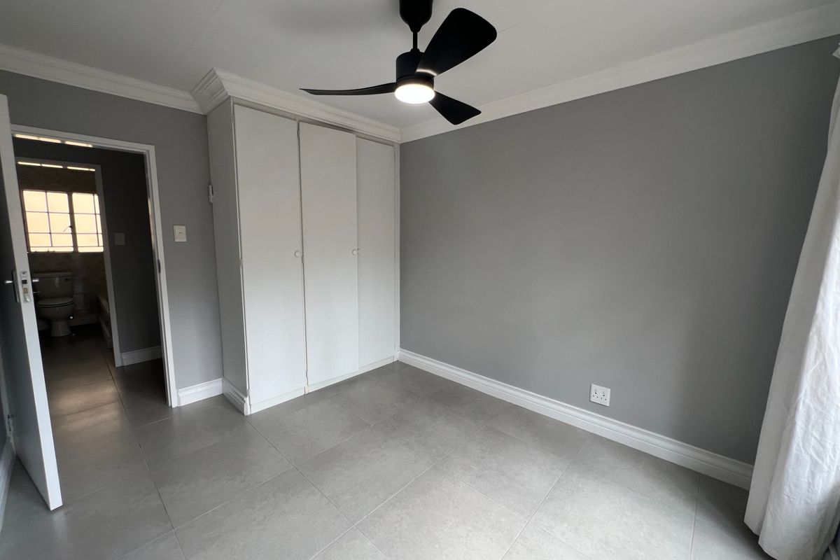 A clean, neutral bedroom featuring built-in cupboards, modern grey floor tiles, and a contemporary ceiling fan, with an adjacent bathroom visible through the doorway.