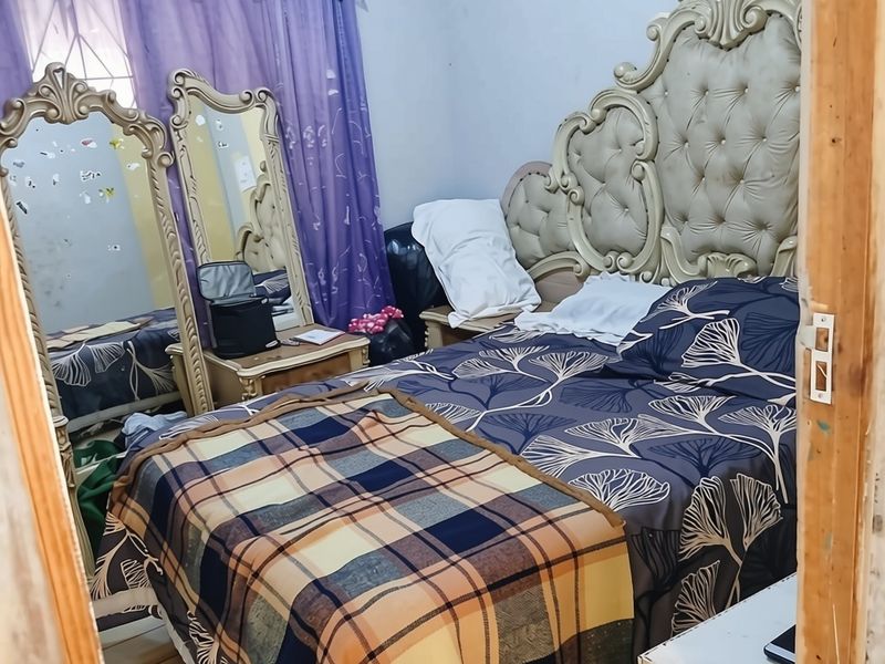 A bedroom featuring a window with curtains and a wooden door frame, showing signs of wear on the walls and trim.