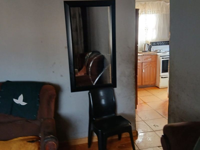 Open plan living area leading into a kitchen with tiled floors and built-in cabinetry, showing signs of wear and wall damage.