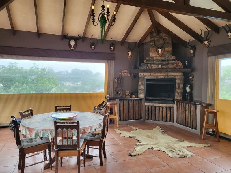 Spacious covered patio featuring exposed timber rafters, a stone-clad fireplace, built-in cabinetry, and large retractable blinds offering scenic views.