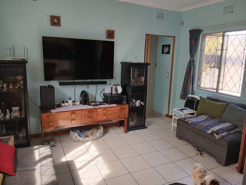 A living room featuring light blue walls, tiled flooring, and a window with security bars, providing a functional and well-lit living space.