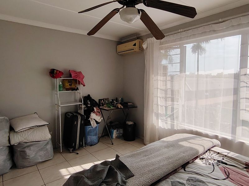 A bedroom featuring tiled flooring, a ceiling fan, and a wall-mounted air conditioning unit, with large windows fitted with security bars.