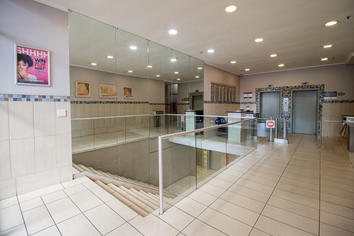 A modern commercial building lobby featuring tiled flooring, glass balustrades, security turnstiles, and elevator access.