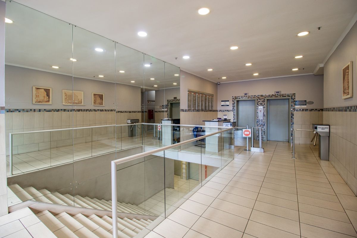 A professional commercial building lobby featuring tiled floors, a glass balustrade, mirrored walls, security turnstiles, and elevator access.